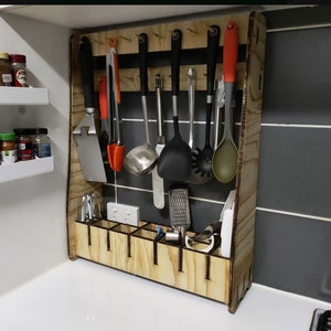 Könnte beinhalten: Ein Küchenutensilien-Organizer aus Holz mit einem Gestell zum Aufhängen von Utensilien und einem Fach zur Aufbewahrung kleiner Gegenstände. Der Organizer ist an einer Wand montiert und hat eine helle Holzoptik.