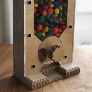 May include: A wooden candy dispenser with a clear acrylic front panel. The dispenser is filled with colorful candies and has a crank on the side to dispense the candy.