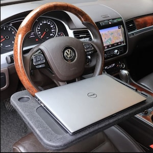 May include: A black car steering wheel with a wooden rim and a silver laptop on a black tray. The laptop has the Dell logo. The tray is attached to the car seat, creating a mobile workspace.