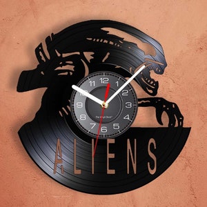 May include: A black vinyl record clock featuring a silhouette of an alien creature from the movie "Aliens". The clock face is white with black numbers and hands. The words "The Vinyl Clock" and "ALIENS" are printed on the record.