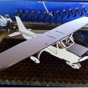 May include: A white wooden model airplane being cut by a laser cutter. The airplane is sitting on a honeycomb-shaped surface.