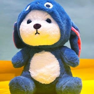 May include: A blue plush toy with a white face and a blue hooded costume. The hood has a white eye and a small white tooth on the front.