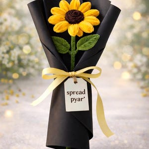 Puede incluir: Un ramo de girasoles hecho a mano envuelto en papel negro y atado con una cinta amarilla. El girasol tiene pétalos amarillos y un centro marrón. Una etiqueta dice "spread pyar".