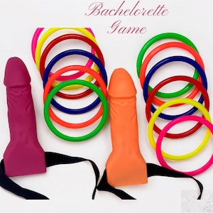 May include: A bachelorette party game set featuring two phallic-shaped objects in pink and orange, with black ribbons attached. Colorful rings in red, green, yellow, blue, and pink are arranged around the objects. The text "Bachelorette Game" is written in cursive.