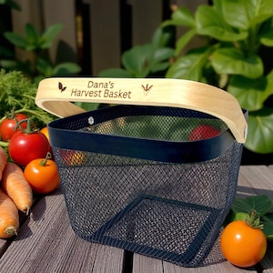 May include: A black mesh harvest basket with a wooden handle. The handle has the text "Dana's Harvest Basket". The basket is filled with tomatoes and carrots. The basket is sitting on a wooden surface.