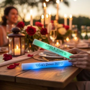 May include: Two glowing LED foam wands with personalized text. One is green and reads "Linda & Vitaly 07/05/2025", the other is blue and says "Eva's Quince 2025". The wands are on a wooden table, with a blurred background of a party.