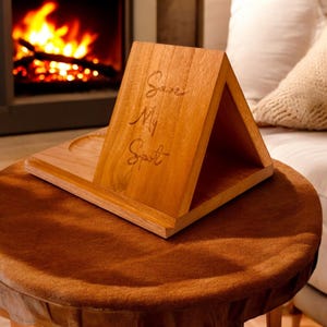 May include: Wooden desk organizer with a triangular design and the engraved text "Save My Spot." The organizer is made of light brown wood and sits on a round, brown table. A fireplace with a burning fire is visible in the background.