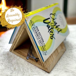 May include: A wooden book stand with a triangular shape, holding a book titled "Eight Dates" by John Gottman, PhD and Julie Schwartz Gottman, PhD. The stand is made of light brown wood and has a small shelf for glasses.