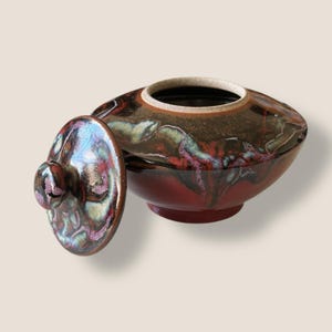 May include: A ceramic decorative bowl with a lid. The bowl is a deep red color with a dark brown and iridescent glaze. The lid is off to the side, revealing the interior of the bowl.