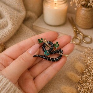 May include: A black and gold brooch in the shape of a snake with green leaf accents and a green gem. The brooch is a piece of jewellery, likely for a jacket or scarf. The snake is coiled and detailed.