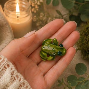 May include: A green frog brooch with emerald-coloured detailing. The frog has a glossy finish and is decorated with small, sparkling crystals. The brooch is held in a hand, with a lit candle and greenery in the background.
