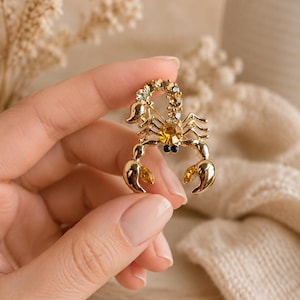 May include: A gold-toned scorpion brooch featuring a central amber-coloured crystal, smaller clear crystals along the curved tail, and two amber-coloured marquise-cut crystals set within the pincers, with two small black beads representing the eyes.