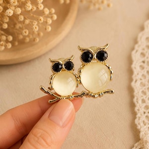 May include: A gold-toned metal brooch featuring two owls perched on a branch. Each owl has a round, translucent cream-colored stone for a body and two small, circular black stones for eyes. The metal frame has a polished finish.