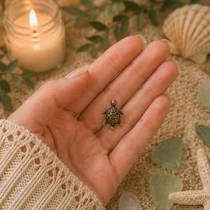 May include: A miniature bronze turtle charm, held in a hand. The charm features a detailed shell pattern and is likely a pendant. A lit candle and seashells are visible in the background. The charm is a decorative item.