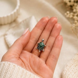 May include: A small, silver turtle pendant with a green enamel shell. The turtle has detailed legs and head. The pendant is held in a hand against a soft, neutral background.