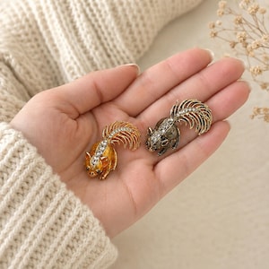 May include: Two squirrel-shaped brooches featuring metallic finishes and clear crystal accents. One brooch is finished in a warm gold tone, while the other features a darker, antique-style metallic finish. Both pieces display detailed tails.