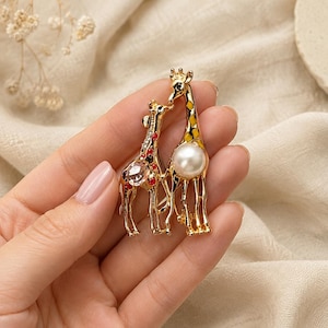 Peut inclure: Une broche en métal doré représentant deux girafes côte à côte. La plus petite girafe est ornée de cristaux rouges et transparents, tandis que la plus grande présente des taches en émail jaune et une perle blanche artificielle sur le corps.