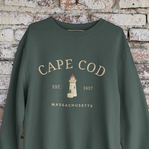 Puede incluir: Una sudadera verde oscuro con un estampado gráfico blanco de un faro y el texto "CAPE COD EST. 1637 MASSACHUSETTS".