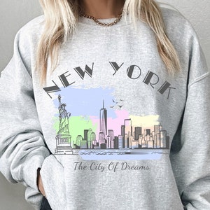 Könnte beinhalten: Grauer Pullover mit einem grafischen Druck der New Yorker Skyline, einschließlich der Freiheitsstatue und des Empire State Building. Der Text "NEW YORK" ist in großer Schrift über der Skyline gedruckt, und der Text "The City Of Dreams" ist unter der Skyline gedruckt.