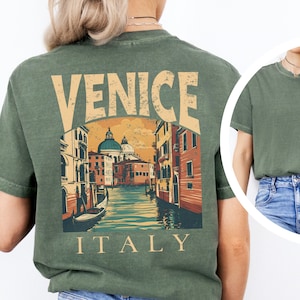 May include: An olive green t-shirt featuring a large graphic on the back of a Venice canal scene with the text VENICE ITALY in a distressed font. The front left chest area displays a smaller graphic of a gondola with the text VENICE ITALY.