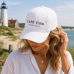 May include: A white baseball cap featuring the dark blue embroidered text CAPE COD MASSACHUSETTS on the front panel. The cap has a structured crown, a curved brim, and eyelets for ventilation, constructed from a solid white fabric material.