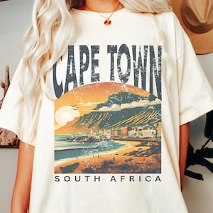 May include: A white t-shirt featuring a vintage-style graphic of Cape Town, South Africa. The graphic depicts a sunset over the city with Table Mountain in the background. The text "Cape Town" is in large, bold letters above the graphic, and the text "South Africa" is below the graphic.