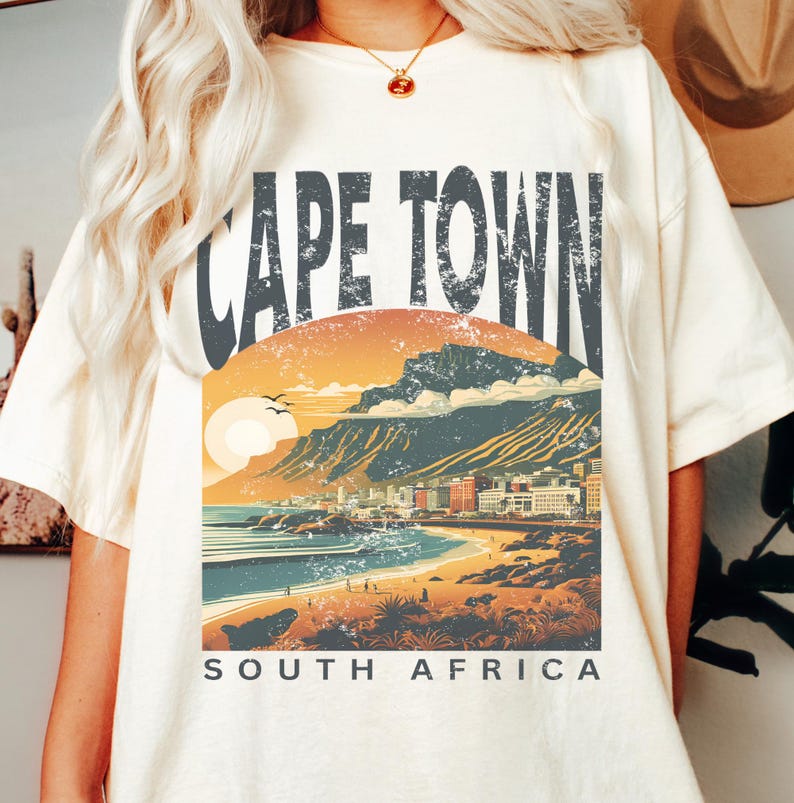 May include: A white t-shirt featuring a vintage-style graphic of Cape Town, South Africa. The graphic depicts a sunset over the city with Table Mountain in the background. The text "Cape Town" is in large, bold letters above the graphic, and the text "South Africa" is below the graphic.