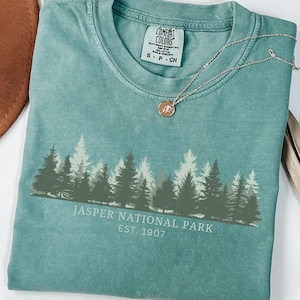 May include: A teal short-sleeved t-shirt featuring a graphic of a forest silhouette in shades of green and white. The text reads "JASPER NATIONAL PARK EST. 1907". A thin gold-toned chain necklace with a small circular pendant rests on the neckline.