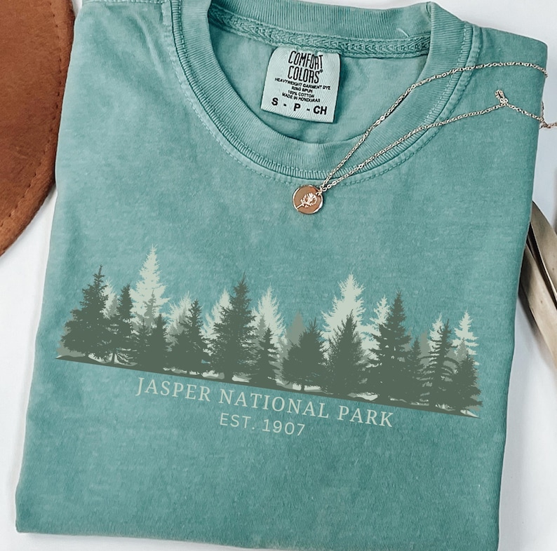 May include: A teal short-sleeved t-shirt featuring a graphic of a forest silhouette in shades of green and white. The text reads "JASPER NATIONAL PARK EST. 1907". A thin gold-toned chain necklace with a small circular pendant rests on the neckline.