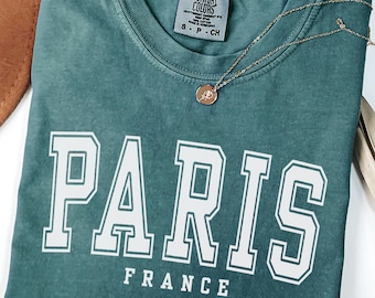 May include: A teal crewneck t-shirt with a vintage-wash finish featuring the white text PARIS FRANCE in a collegiate font. The shirt has a Comfort Colors brand label at the inner neckline and is paired with a delicate gold-toned chain necklace.