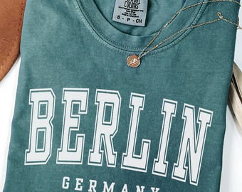 Pode incluir: Uma camiseta Comfort Colors verde-azulado com a palavra "BERLIN" em letras brancas e "GERMANY" em baixo. Um colar de ouro com um pequeno pingente repousa sobre a camisa. A camisa está dobrada cuidadosamente sobre uma superfície branca.