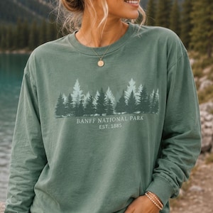 May include: A sage green long-sleeve crewneck sweatshirt featuring a graphic print of a forest of pine trees in varying shades of green. Below the trees, the text reads BANFF NATIONAL PARK EST. 1885 in a clean, white, sans-serif font.