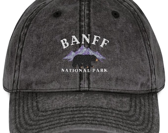 May include: A distressed black baseball cap with the word "BANFF" embroidered in white above a mountain and bear graphic. The text "NATIONAL PARK" is below the graphic. The cap has a curved brim and an adjustable strap.