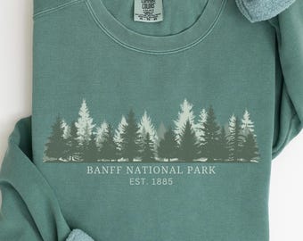 May include: A teal crewneck sweatshirt featuring a graphic of a forest silhouette in varying shades of green. Below the trees, the text reads BANFF NATIONAL PARK EST. 1885 in white. The interior neck label displays the Comfort Colors brand name.