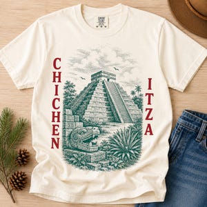 May include: A cream-coloured short-sleeve t-shirt featuring a dark green graphic illustration of the Chichen Itza pyramid, a stone serpent head, and tropical plants. The text CHICHEN ITZA is printed in dark red vertically on both sides of the graphic.