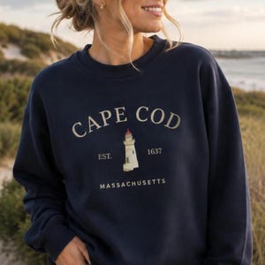 May include: A navy blue crewneck sweatshirt featuring the text CAPE COD in a curved font above a graphic of a lighthouse. The text EST. 1637 is positioned on either side of the lighthouse, with the word MASSACHUSETTS printed below the graphic.