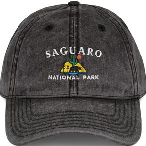 Könnte beinhalten: Eine anthrazitfarbene, säuregewaschene Baseballkappe mit einer Stickerei, die einen Bären, einen Saguaro-Kaktus und eine Sonne über gelben Hügeln zeigt. Der Text SAGUARO NATIONAL PARK ist in Weiß über und unter der Grafik eingestickt.
