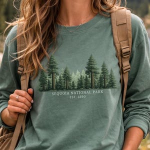 May include: A sage green long-sleeve crewneck shirt featuring a graphic of a forest of sequoia trees. Below the trees, the text reads "SEQUOIA NATIONAL PARK" and "EST. 1890" in a clean, white sans-serif font. The shirt is worn with a tan backpack.