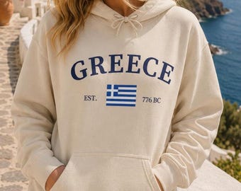 Peut inclure: Un sweat-shirt à capuche couleur crème arborant le mot GREECE en lettres bleu foncé, un graphique du drapeau grec bleu et blanc, et le texte EST. 776 BC. Le vêtement comprend une capuche à cordon de serrage et une poche kangourou frontale.