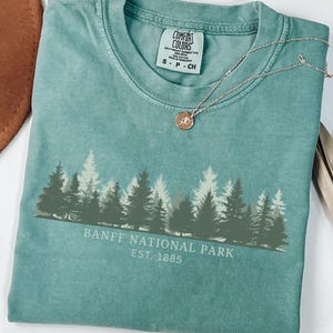 May include: A sage green short-sleeve t-shirt featuring a graphic of a forest silhouette and the text "BANFF NATIONAL PARK EST. 1885" in white. The shirt has a crew neckline and a Comfort Colors brand label on the inside collar showing size S P CH.