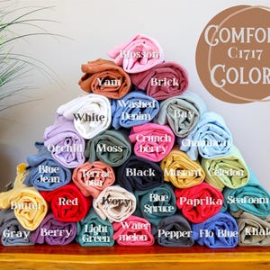 May include: A pyramid of rolled Comfort Colors C1717 t-shirts in various colors including Blossom, Yam, Brick, White, Washed Denim, Bay, Orchid, Moss, Crunchberry, Chambray, Blue Jean, Terracotta, Black, Mustard, Celedon, Butter, Red, Ivory, Blue Spruce, Paprika, Seafoam, Gray, Berry, Light Green, Watermelon, Pepper, Flo Blue, and Khaki.