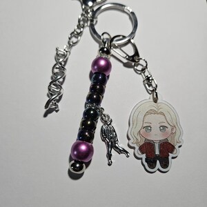 May include: A keychain featuring a silver-toned DNA helix charm, a strand of purple and iridescent dark beads, a small silver figure charm, and an acrylic charm of a blonde character wearing a red jacket, all attached to a silver metal ring.