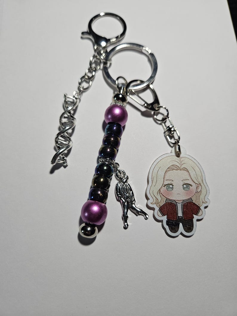 May include: A keychain featuring a silver-toned DNA helix charm, a strand of purple and iridescent dark beads, a small silver figure charm, and an acrylic charm of a blonde character wearing a red jacket, all attached to a silver metal ring.