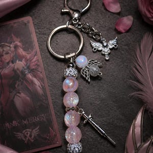 May include: A keychain featuring a series of iridescent pink spherical beads, silver-toned spacers, and a silver-toned metal sword charm. The keychain includes a silver-toned ring and a small wing-shaped charm with clear crystals attached to the chain.