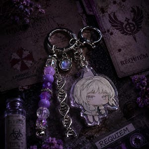 May include: A silver-toned keyring featuring a split ring and lobster clasp, adorned with a strand of purple and clear faceted beads, a silver DNA helix charm, a small round iridescent charm, and a clear acrylic charm of a stylised anime character.
