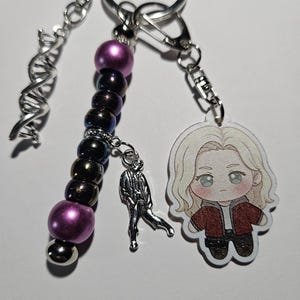 May include: A keychain featuring a silver DNA helix charm, a strand of purple and iridescent dark beads, a small silver figure charm, and an acrylic charm of a blonde character wearing a red jacket, all attached to a silver metal ring.