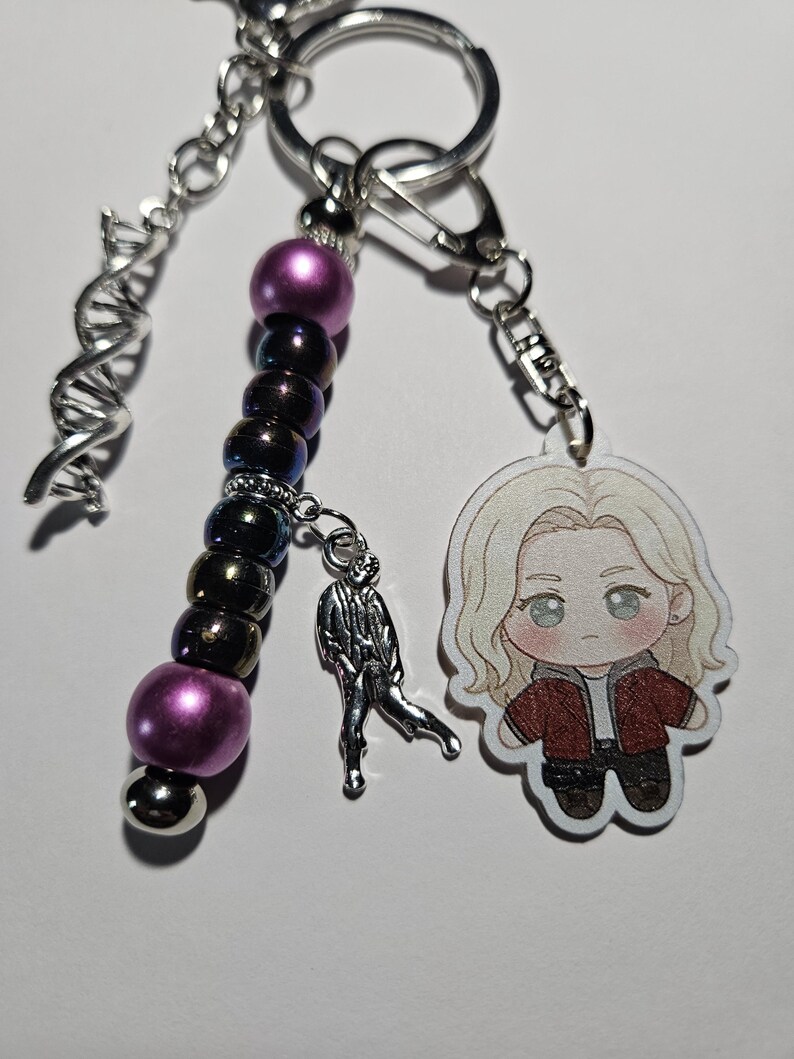May include: A keychain featuring a silver DNA helix charm, a strand of purple and iridescent dark beads, a small silver figure charm, and an acrylic charm of a blonde character wearing a red jacket, all attached to a silver metal ring.