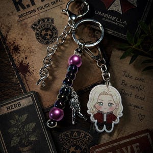 May include: A keychain featuring a silver-toned DNA helix charm, a strand of iridescent dark purple and magenta beads, a small silver figure charm, and a flat acrylic charm depicting a blonde character in a red jacket and black shirt.