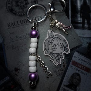 May include: A silver-toned keychain featuring a vertical strand of alternating purple and white beads, a silver DNA helix charm, a small silver figure charm, and a clear acrylic charm depicting a stylized anime-style character with short hair.