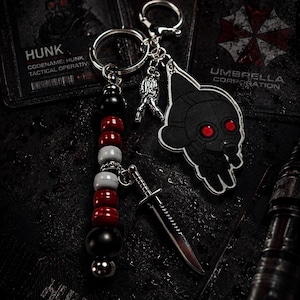 May include: A keychain featuring a character charm with red eyes and a gas mask, a silver-toned dagger charm, and a strand of black, red, and white beads. The keychain includes a metal ring and clasp, with a small figure charm attached to the chain.